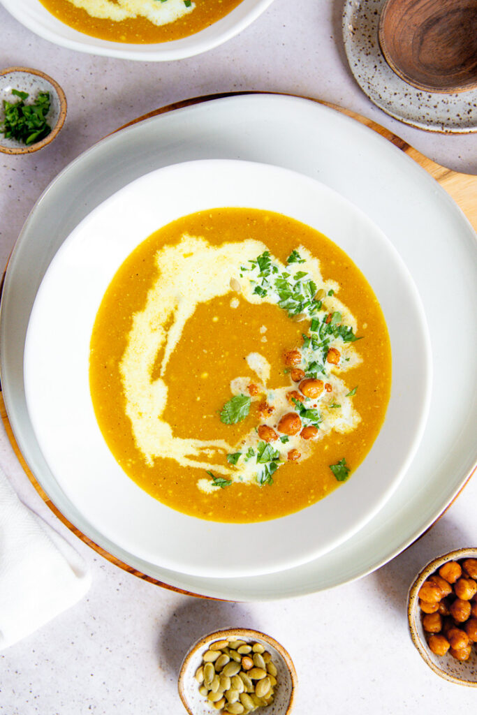 a white bowl on a white plate filled with butternut squash soup topped with a drizzle of coconut milk, diced cilantro, roasted chickpeas, and pepitas surrounded by a few small bowls filled with the toppings