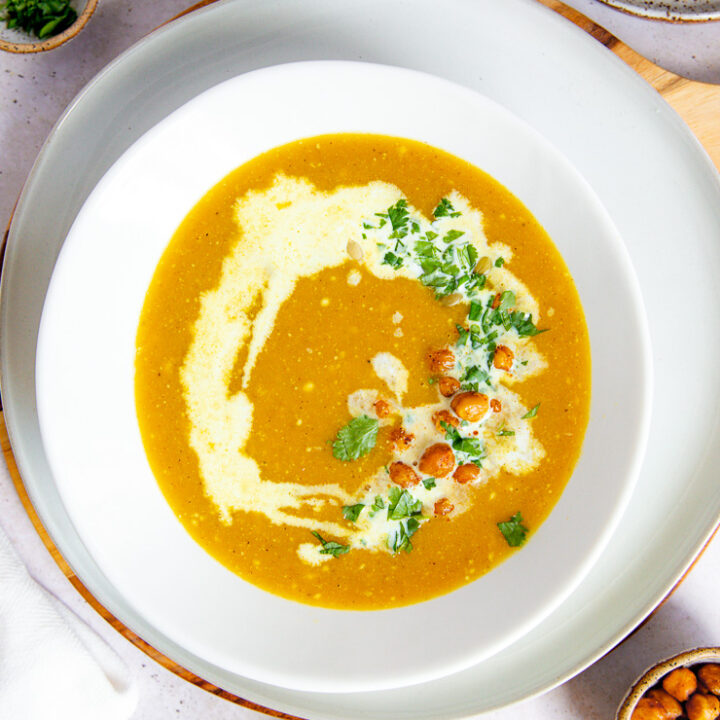 a white bowl on a white plate filled with butternut squash soup topped with a drizzle of coconut milk, diced cilantro, roasted chickpeas, and pepitas surrounded by a few small bowls filled with the toppings