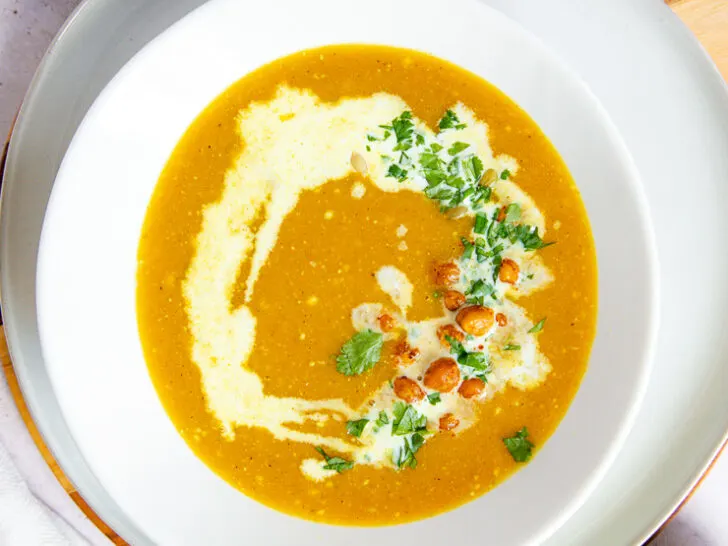 a white bowl on a white plate filled with butternut squash soup topped with a drizzle of coconut milk, diced cilantro, roasted chickpeas, and pepitas surrounded by a few small bowls filled with the toppings