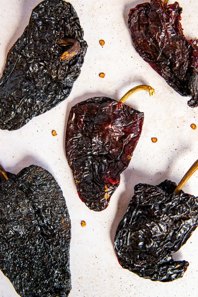 Five ancho chile peppers laying on a light tan surface surrounded by seeds.