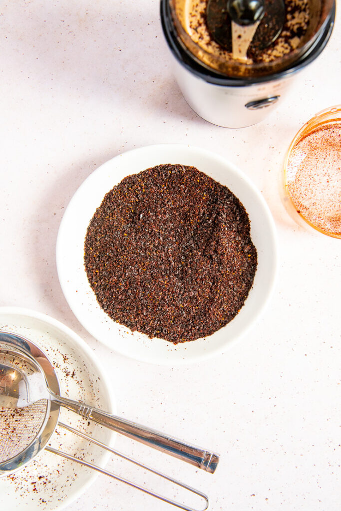 A small white plate with blended ancho chile powder next to a spice grinder and a small mesh sieve.