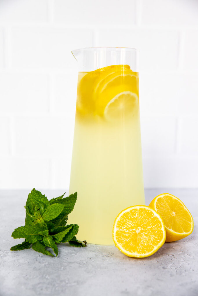 A tall glass pitcher with lemonade and lemon rounds with lemon halves and mint leaves sitting in front of it.