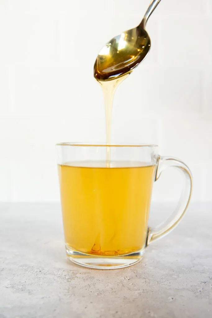 A spoon dripping honey into a clear glass mug with tea.