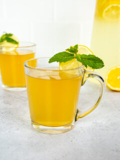 Two glass mugs with medicine ball tea, mint leaves, and lemon rounds next to a pitcher of lemonade.