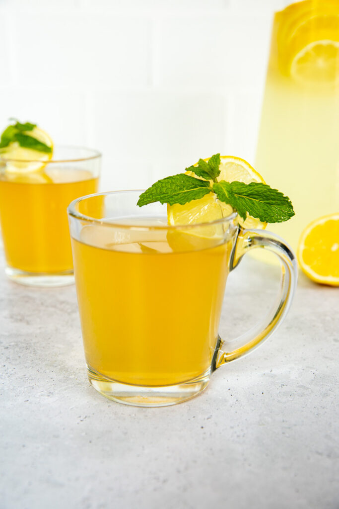 Two glass mugs with medicine ball tea, mint leaves, and lemon rounds next to a pitcher of lemonade.