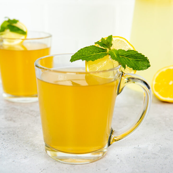 Two glass mugs with medicine ball tea, mint leaves, and lemon rounds next to a pitcher of lemonade.