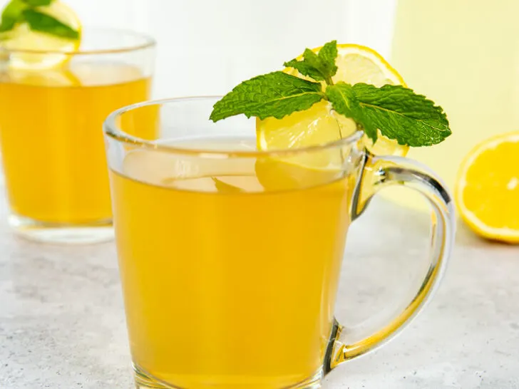 Two glass mugs with medicine ball tea, mint leaves, and lemon rounds next to a pitcher of lemonade.