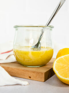 A small Weck glass jar with honey lemon vinaigrette and a spoon in it with a lemon halved sitting next to it.