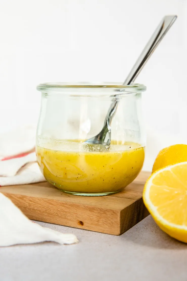 A small Weck glass jar with honey lemon vinaigrette and a spoon in it with a lemon halved sitting next to it.