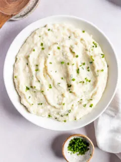 Horseradish Mashed Potatoes with chives in a while bowl next to a wooden spoon and linen napkin.