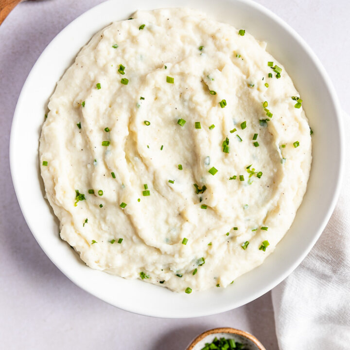 Horseradish Mashed Potatoes with chives in a while bowl next to a wooden spoon and linen napkin.