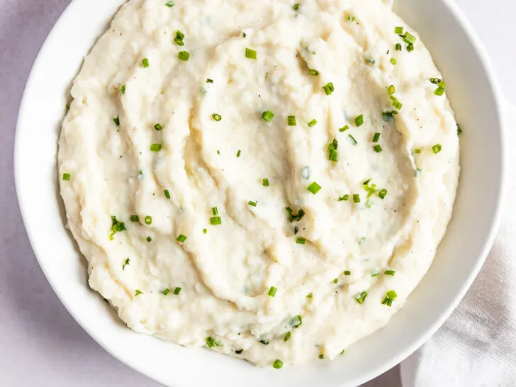 Horseradish Mashed Potatoes with chives in a while bowl next to a wooden spoon and linen napkin.