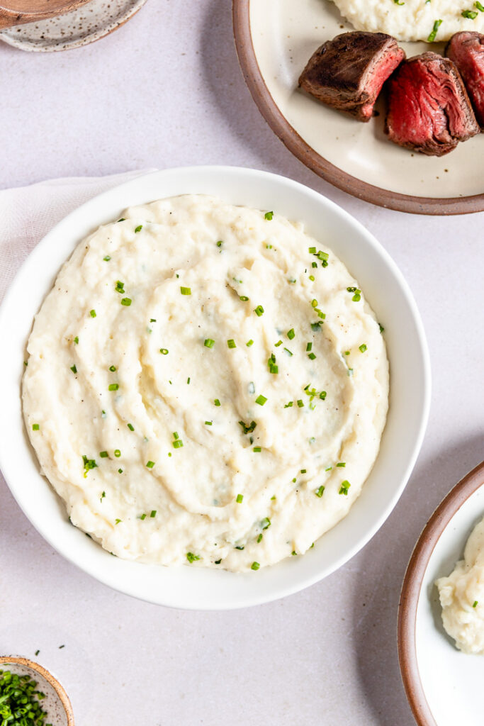 Horseradish Mashed Potatoes in a white bowl topped with chives next to a filled plate with filet mingon slices.