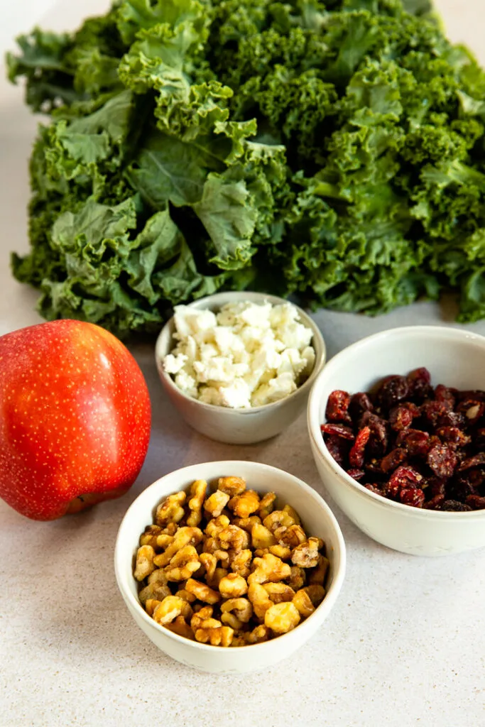 A bunch of kale, red apple, and three small bowls filled with crumbled goat cheese, dried cranberries, and chopped walnuts.