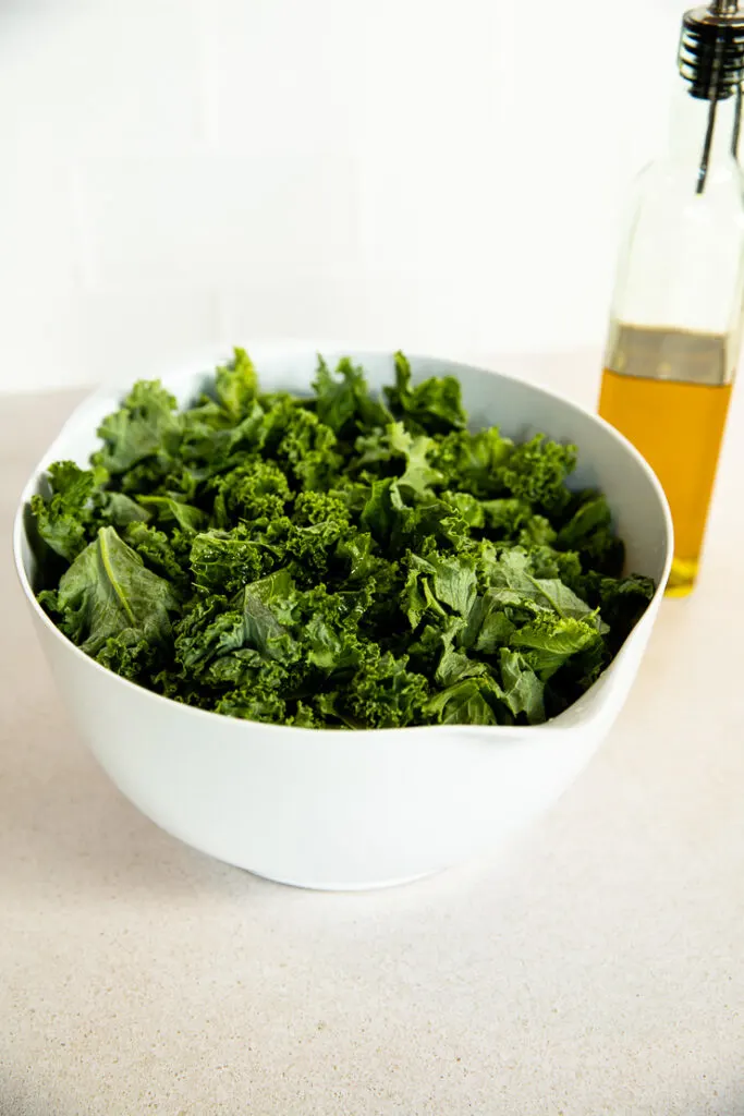 A white mixing bowl full of kale next to a pouring container of olive oil.