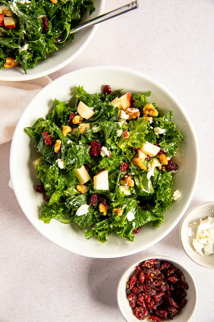 An overhead shot of the Kale Salad with Cranberries next to two small dishes of dried cranberries and goat cheese.