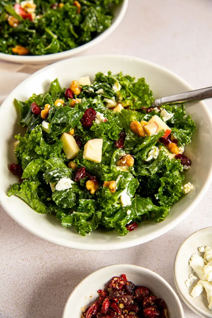 A close up shot of a bowl of salad with a few pieces of chopped apple, walnuts, and dried cranberries poking out of the kale.