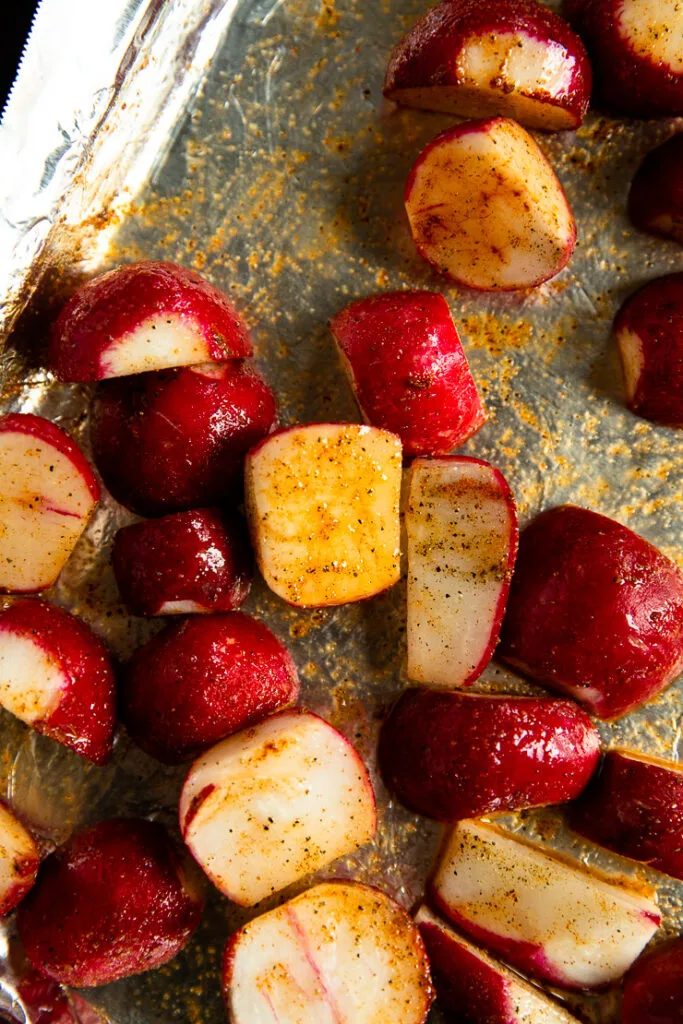 Raw halves of roasted radishes covered in olive oil and seasoning on an aluminum foil covered baking sheet.