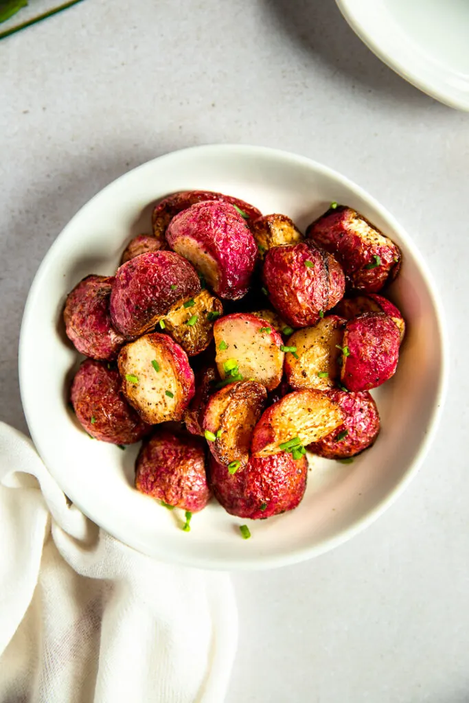 An overhead shot of a white bowl filled with halves of roasted radishes topped with diced chives.