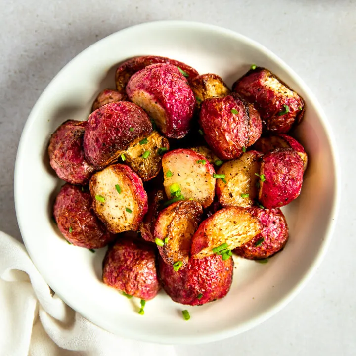 An overhead shot of a white bowl filled with halves of roasted radishes topped with diced chives.