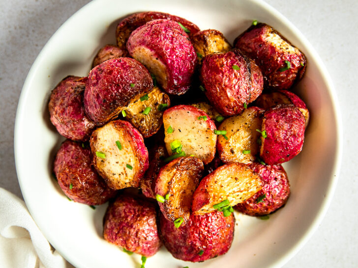 An overhead shot of a white bowl filled with halves of roasted radishes topped with diced chives.