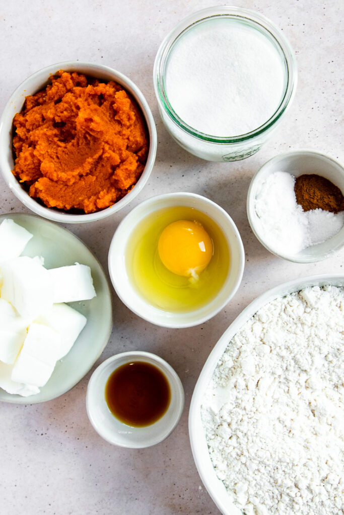 Bowls of varying sizes containing the ingredients to make pumpkin cookies including flour, vanilla extract, crisco cubes, an egg, spices, pumpkin puree, and sugar.