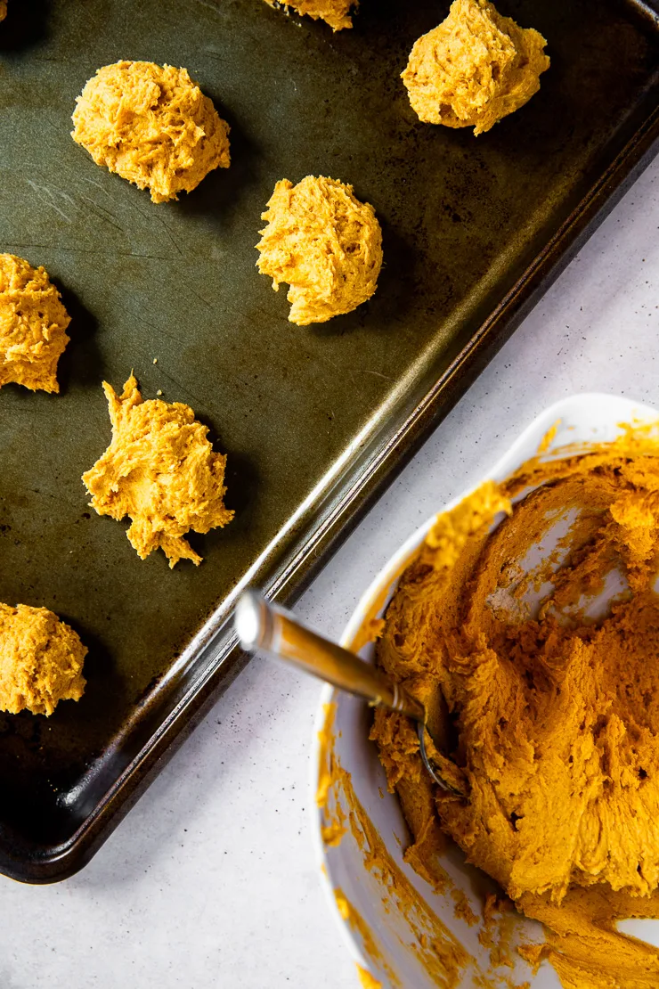 A white bowl of pumpkin cookie dough next to a cookie sheet with scooped dough balls.