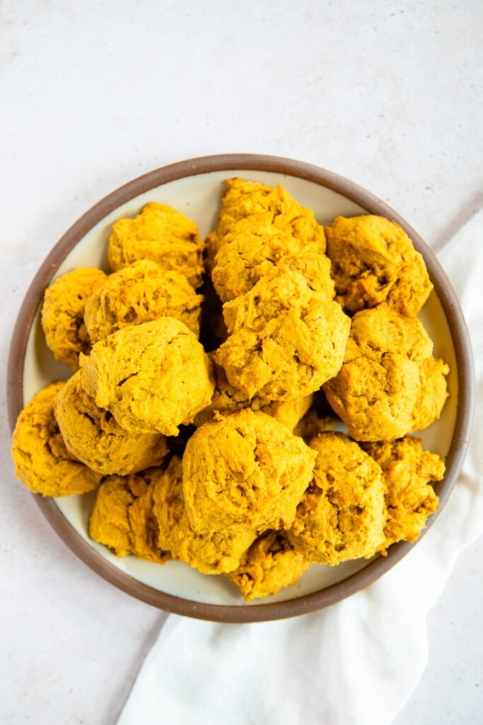 An overhead shot of a pile of soft pumpkin cookies on a plate with a brown rim on a white linen napkin.