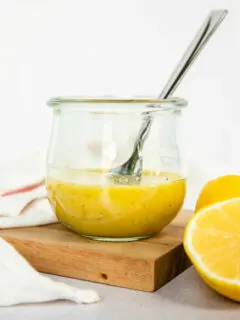 A small Weck glass jar with honey lemon vinaigrette and a spoon in it with a lemon halved sitting next to it.