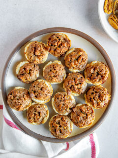 A plate full of pecan tassies on a linen napkin next to a white plate of pecan halves.