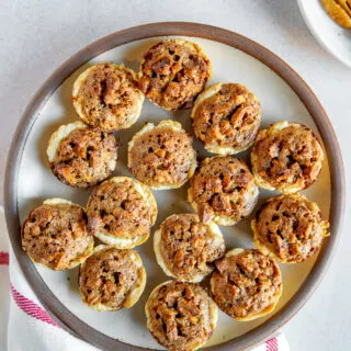A plate full of pecan tassies on a linen napkin next to a white plate of pecan halves.