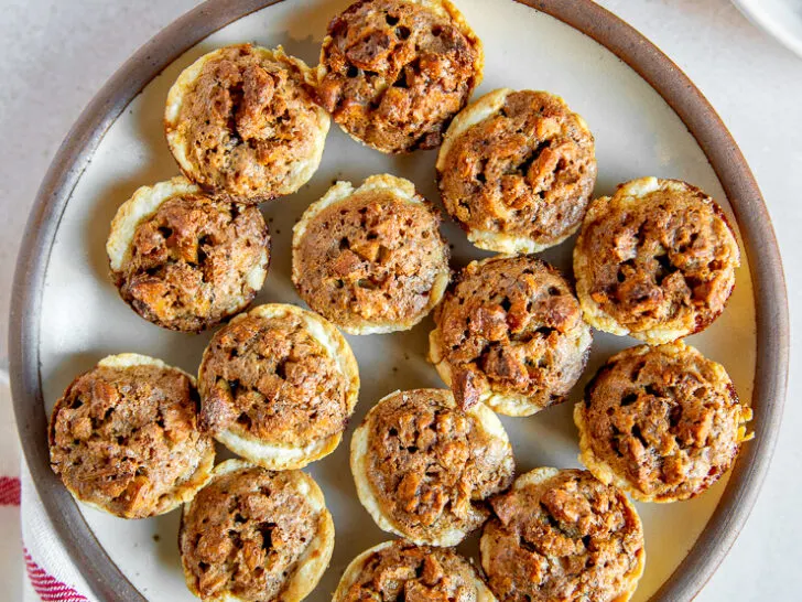 A plate full of pecan tassies on a linen napkin next to a white plate of pecan halves.