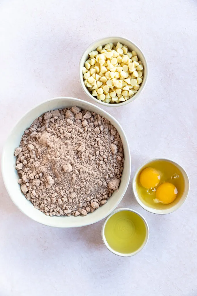Four white bowls of varying sizes with white chocolate chips, red velvet cake mix, two eggs, and vegetable oil in each.