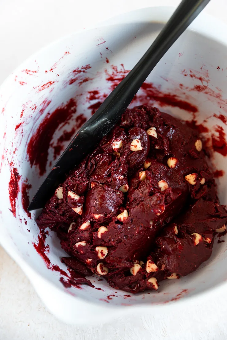 A black spatula stirring red velvet cake mix cookie dough in a big white plastic bowl.
