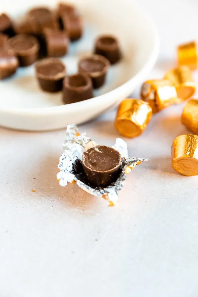 An unwrapped Rolo chocolate still sitting on the gold foil next to two piles of candies, some unwrapped and some still wrapped.