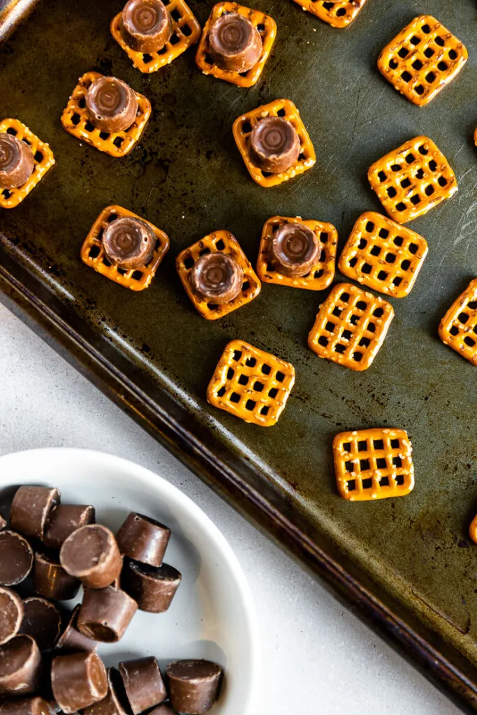 A cookie sheet with square pretzels and Rolos on top next to a small bowl of unwrapped Rolos.