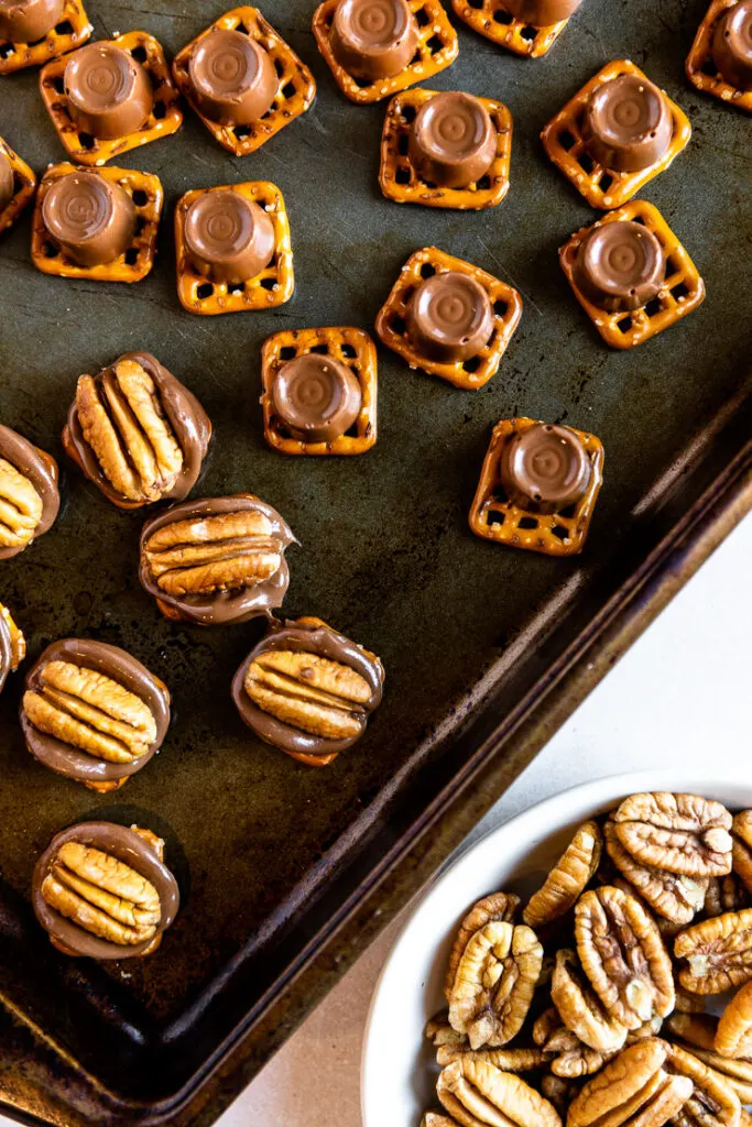 A cookie sheet with melted Rolos on square pretzels and a pecan half topping each one.