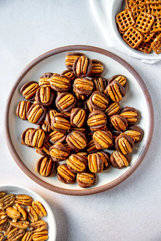 A plate full of rolo pretzel turtles next to two shallow bowls each filled with square pretzels and pecan halves respectively.