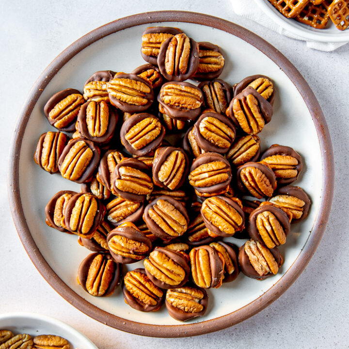 A plate full of rolo pretzel turtles next to two shallow bowls each filled with square pretzels and pecan halves respectively.