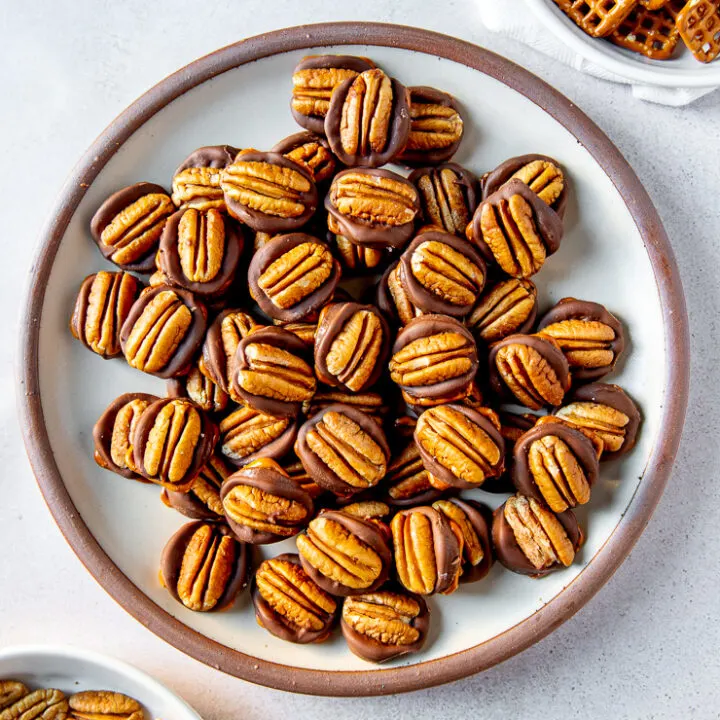 A plate full of rolo pretzel turtles next to two shallow bowls each filled with square pretzels and pecan halves respectively.