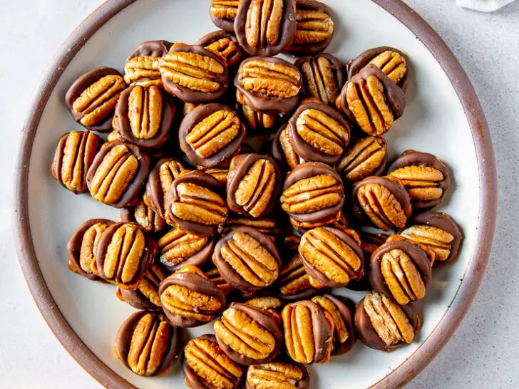 A plate full of rolo pretzel turtles next to two shallow bowls each filled with square pretzels and pecan halves respectively.