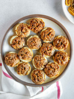 A plate full of pecan tassies on a linen napkin next to a white plate of pecan halves.