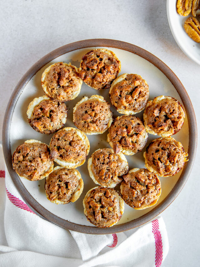 A plate full of pecan tassies on a linen napkin next to a white plate of pecan halves.