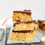 Three stacked scotcharoos on parchment paper in front of a white background with a text overlay that says "The Ultimate Scotcharoos - @theculinarycompass."