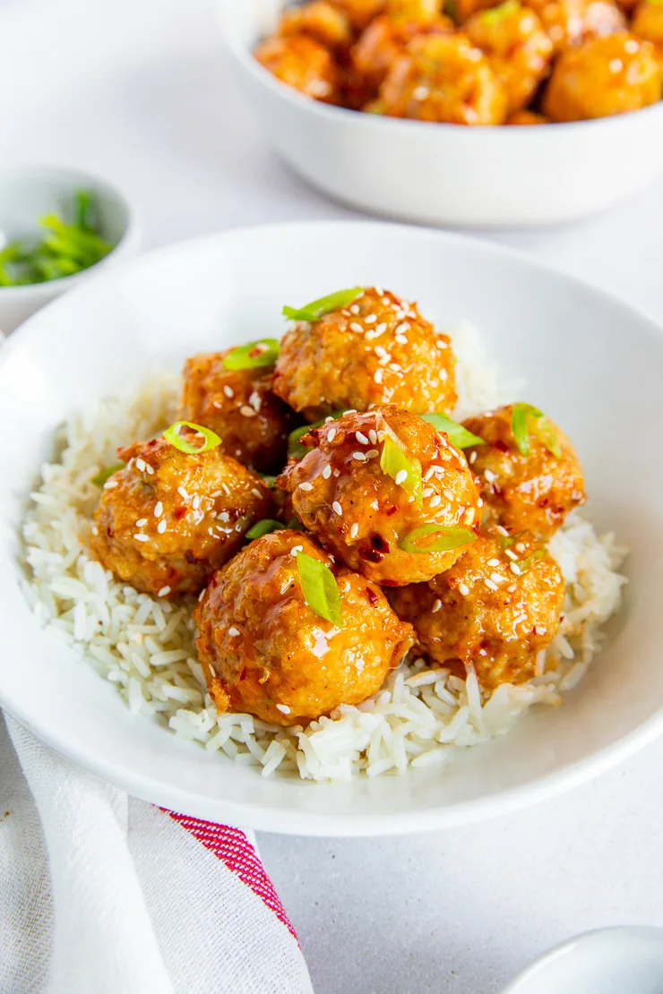 A side angle of a bowl of firecracker meatballs in a white bowl on white rice and green onions on top.