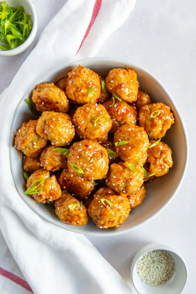 A big white bowl holding a pile of firecracker meatballs covered in sauce, white sesame seeds, and cut green onions.