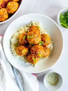 A bowl of white rice and firecracker meatballs with sesame seeds and green onions on top and bowls of each ingredient surrounding it.