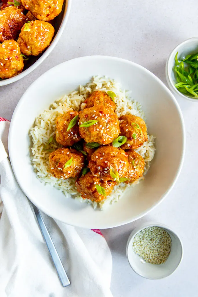 A bowl of white rice and firecracker meatballs with sesame seeds and green onions on top and bowls of each ingredient surrounding it.