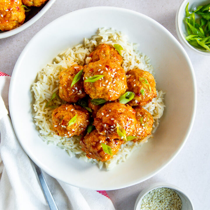 A bowl of white rice and firecracker meatballs with sesame seeds and green onions on top and bowls of each ingredient surrounding it.