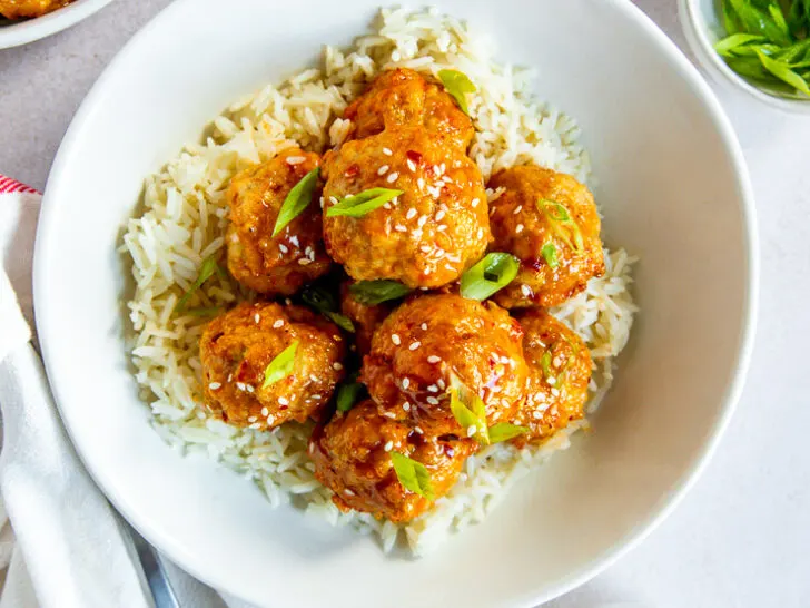 A bowl of white rice and firecracker meatballs with sesame seeds and green onions on top and bowls of each ingredient surrounding it.